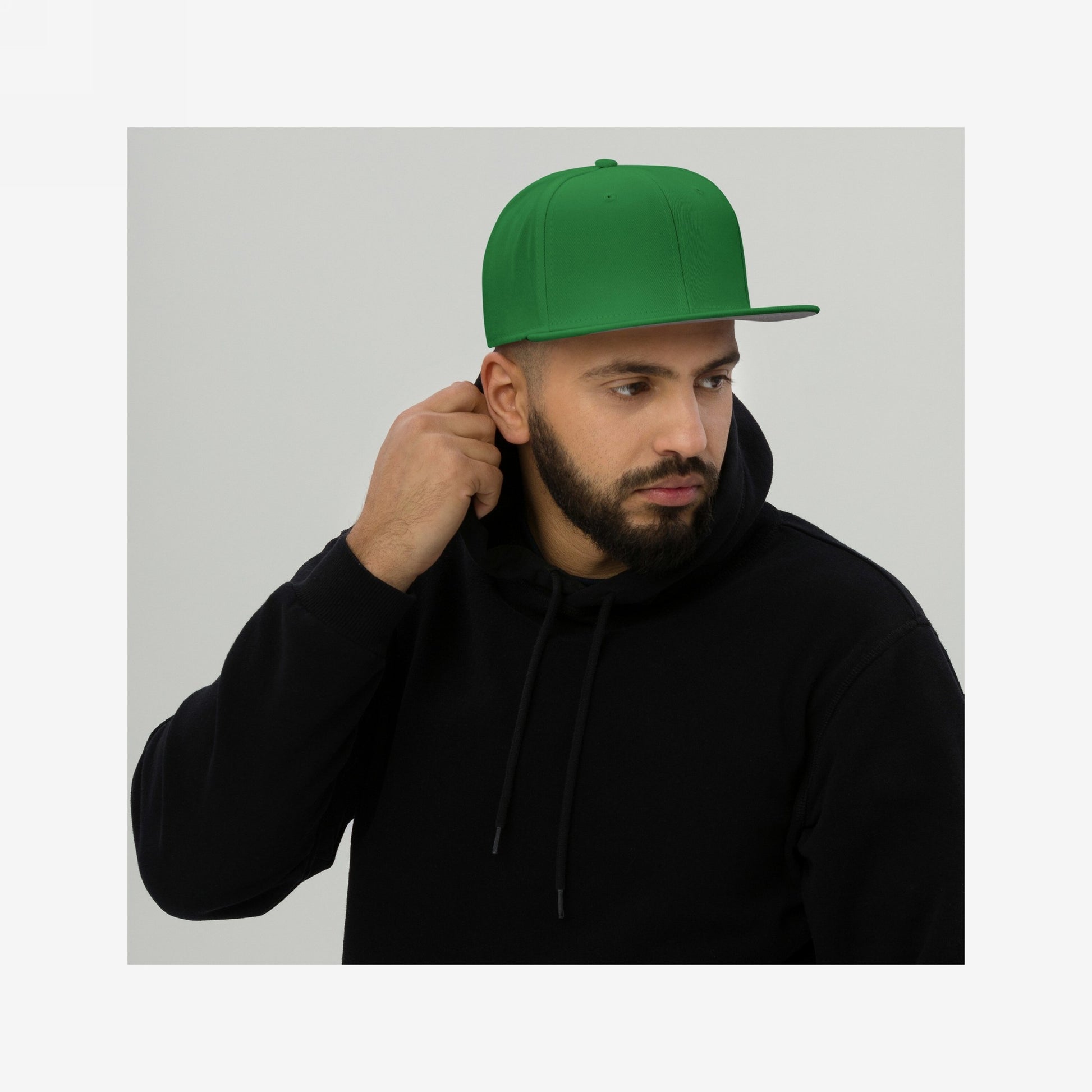 Bearded man wearing a Snapback Hat In Custom Colors, featuring a high-profile crown and flat brim, grasping his black hoodie's hood, against a light-gray background, embodying activist spirit and bold expression.