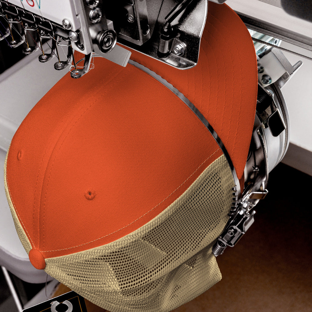 Trucker Hat In Custom Colors being embroidered on an industrial machine, showcasing bright orange panels and tan mesh back, perfect for making bold statements with personalized designs.