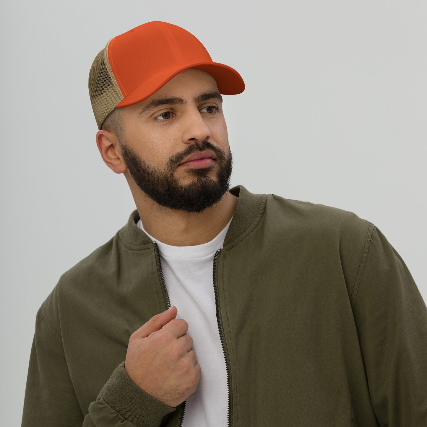 Bearded man models Trucker Hat In Custom Colors, featuring an orange front and tan mesh back, paired with an olive-green jacket. The cap exemplifies expressive, custom embroidery for activism and personal statements.