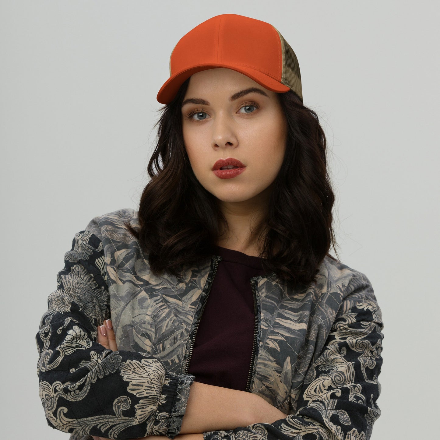 Young woman models the Trucker Hat In Custom Colors with an orange front and mesh back, embodying bold style and activism. She's wearing a patterned jacket, arms crossed, looking directly at the camera.