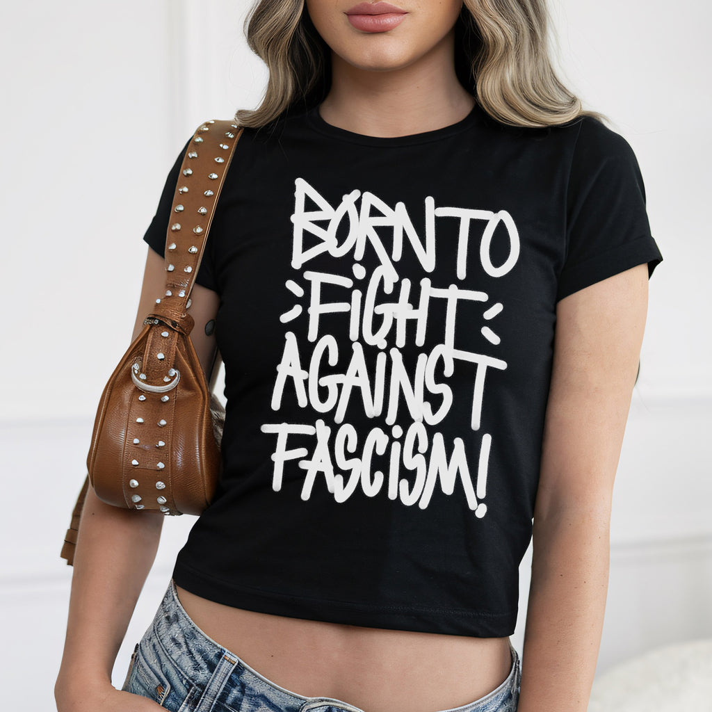 Born To Fight Against Fascism Graffiti Women's Baby Tee, featuring bold white lettering on black fabric, worn with light-wash jeans. The cropped design radiates defiance, epitomizing anti-fascist sentiment and empowerment.