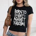 Born To Fight Against Fascism Graffiti Women's Baby Tee, featuring bold white lettering on black fabric, worn with light-wash jeans. The cropped design radiates defiance, epitomizing anti-fascist sentiment and empowerment.