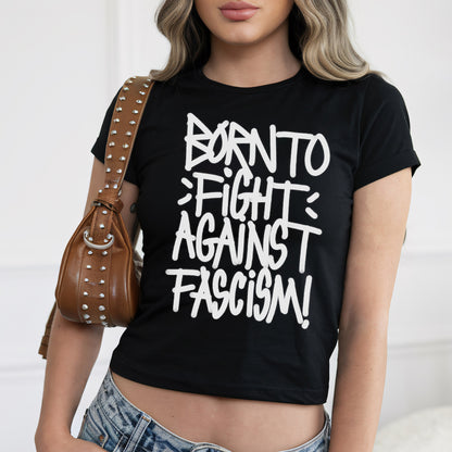 Born To Fight Against Fascism Graffiti Women's Baby Tee, featuring bold white lettering on black fabric, worn with light-wash jeans. The cropped design radiates defiance, epitomizing anti-fascist sentiment and empowerment.