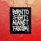 Born To Fight Against Fascism Graffiti Poster displays bold, white graffiti text on a black background, mounted on a textured red wall. Signature tag in the corner, embodying defiance and street protest energy.