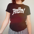 Person wearing a Boston Handstyle Women's Baby Tee featuring bold white graffiti-style text with a halo above the O, paired with black-and-white checkered pants, reflecting Boston's vibrant and bold spirit.