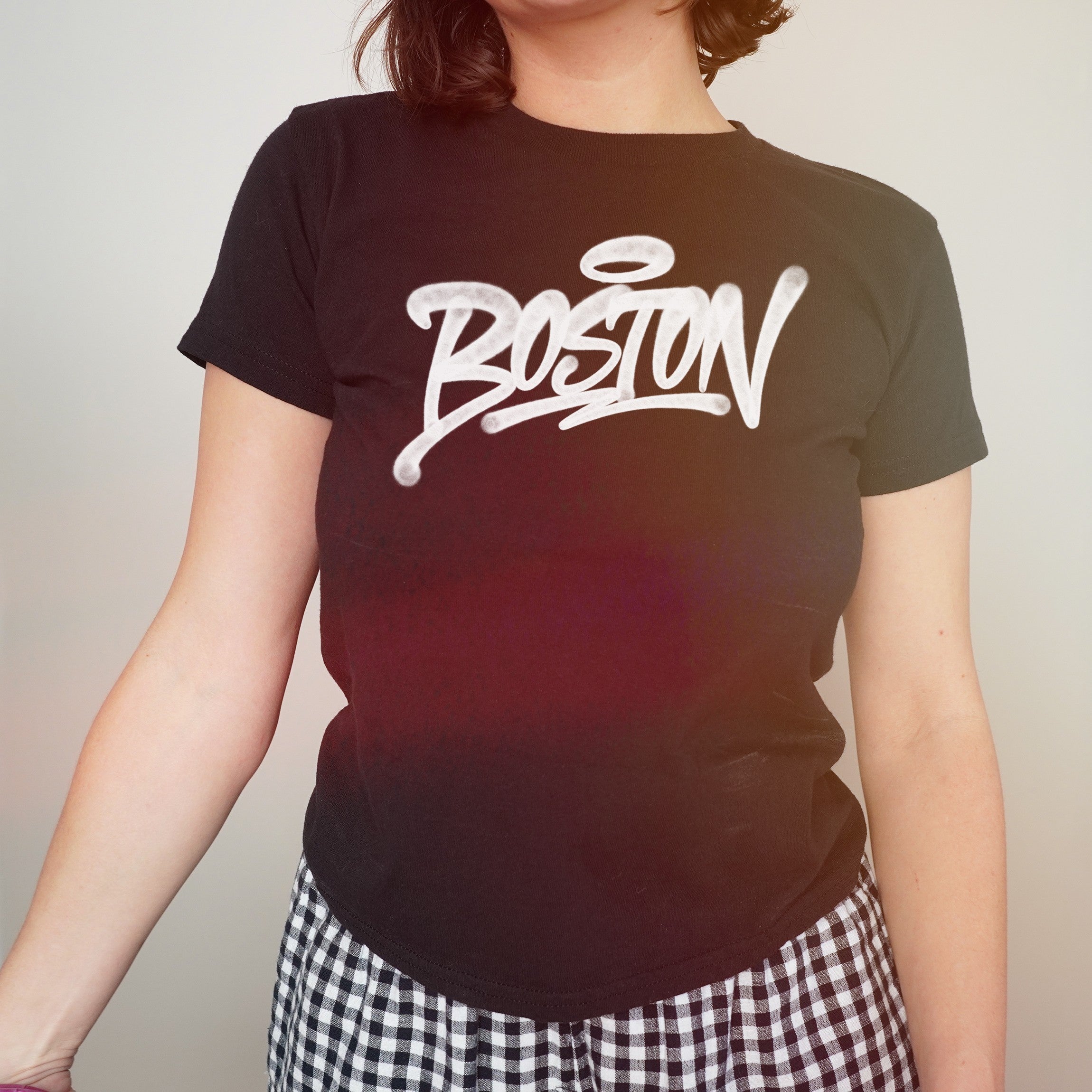 Person wearing a Boston Handstyle Women's Baby Tee featuring bold white graffiti-style text with a halo above the O, paired with black-and-white checkered pants, reflecting Boston's vibrant and bold spirit.
