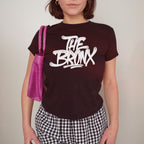 Person wearing the Bronx Handstyle Women's Baby Tee with THE BRONX graffiti-style print, paired with a small purple shoulder bag and black-and-white gingham pants, embodying bold NYC street style.