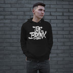 Young man in front of brick wall wearing Bronx Handstyle Hoodie with bold graffiti-style The Bronx graphic, embodying urban style and borough pride. Hands in pocket, looking right.