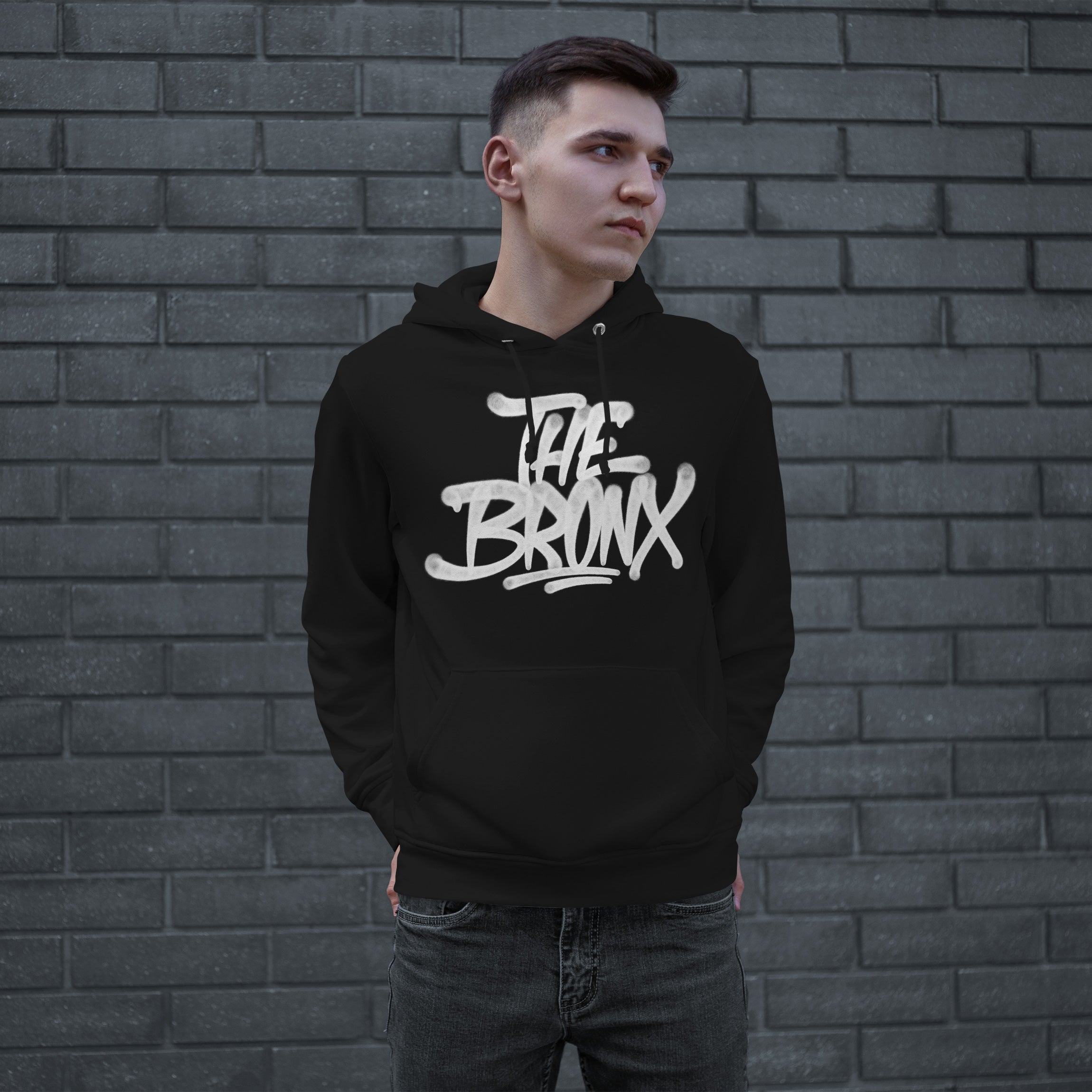 Young man in front of brick wall wearing Bronx Handstyle Hoodie with bold graffiti-style The Bronx graphic, embodying urban style and borough pride. Hands in pocket, looking right.