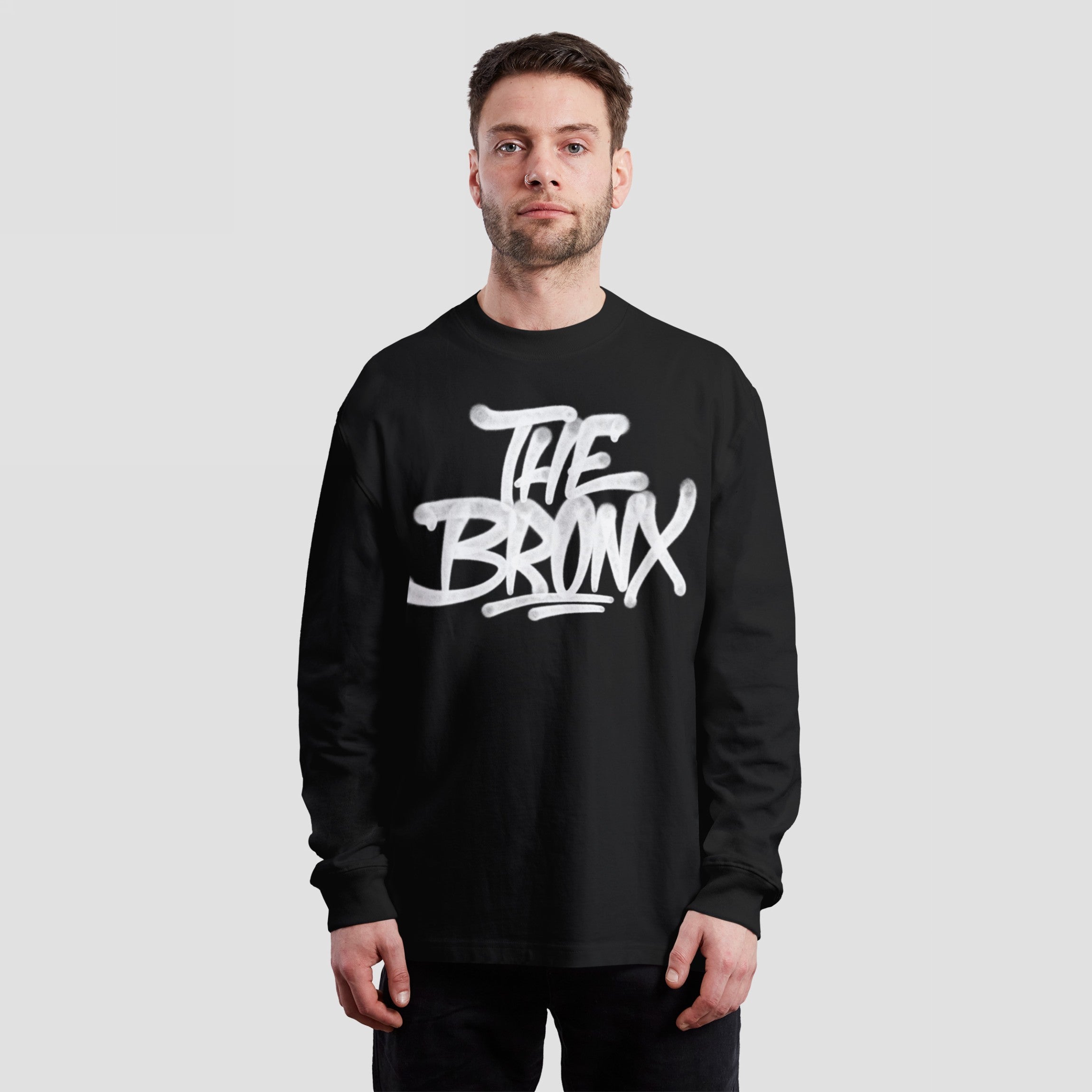 Young man in a black Bronx Handstyle T-shirt with graffiti-style white lettering THE BRONX, embodying NYC pride and rebellious spirit, stands confidently against a light-gray background.