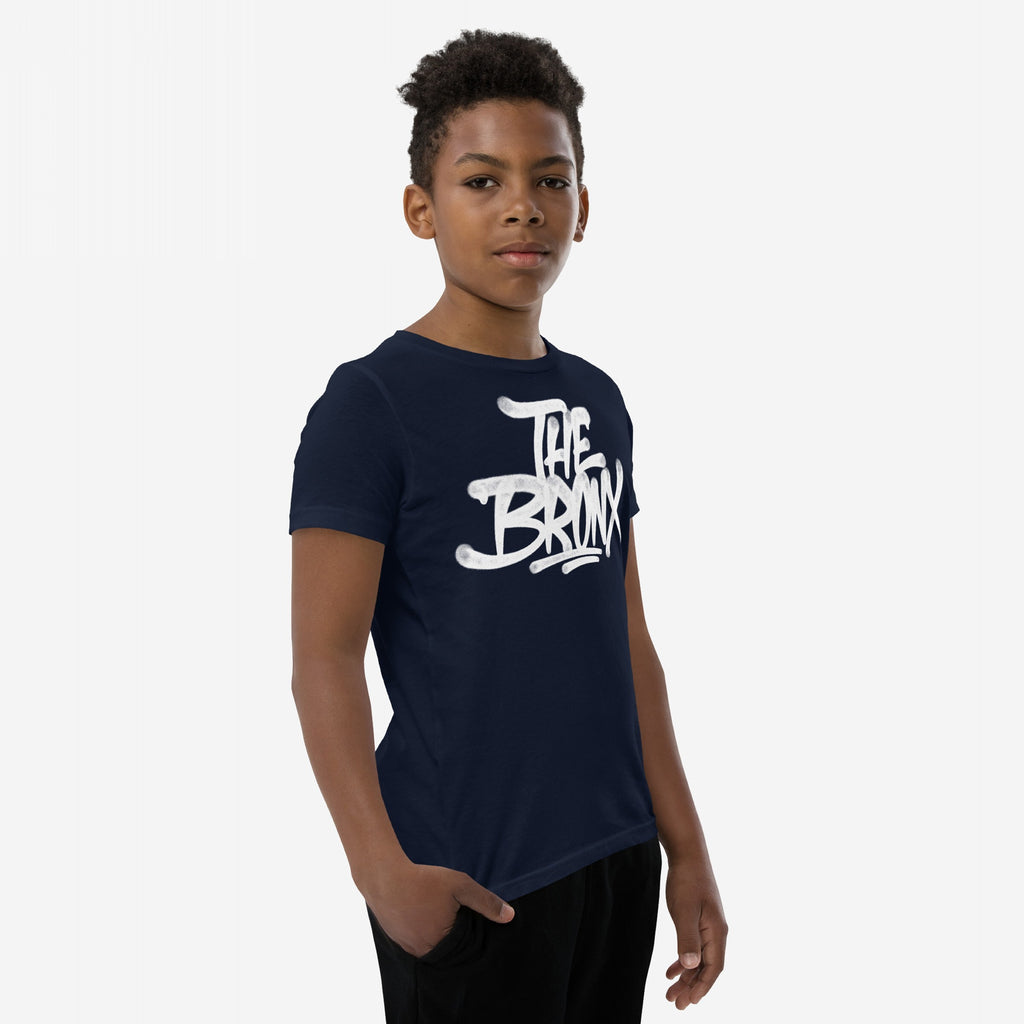 Young boy in a navy Bronx Handstyle T-shirt featuring white graffiti-style The Bronx print, standing with one hand in his pocket against a plain white backdrop.