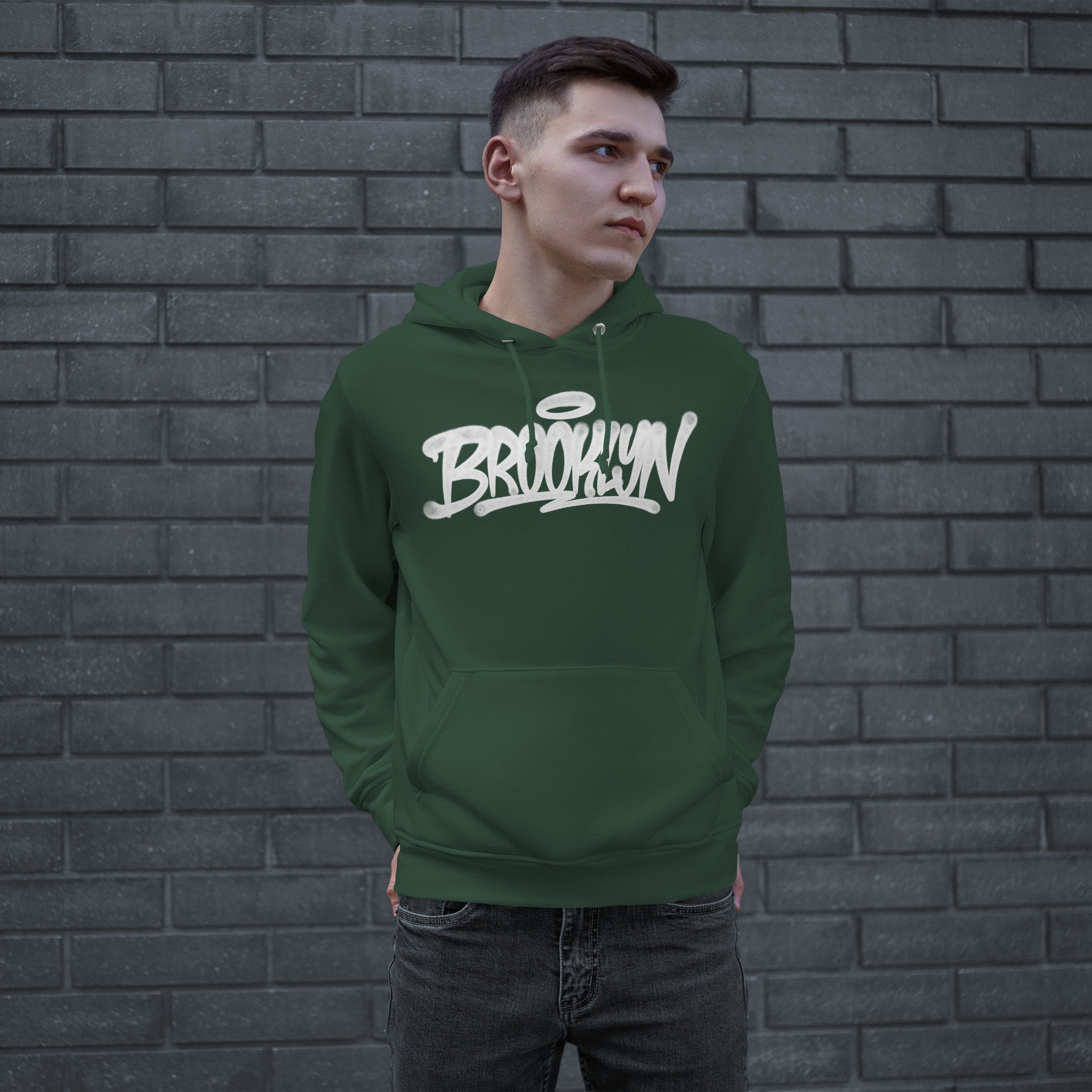 Young man modeling the Brooklyn Handstyle Hoodie, featuring a white graffiti-style BROOKLYN logo with a halo on a dark green pullover. He's standing casually with hands in the kangaroo pocket.