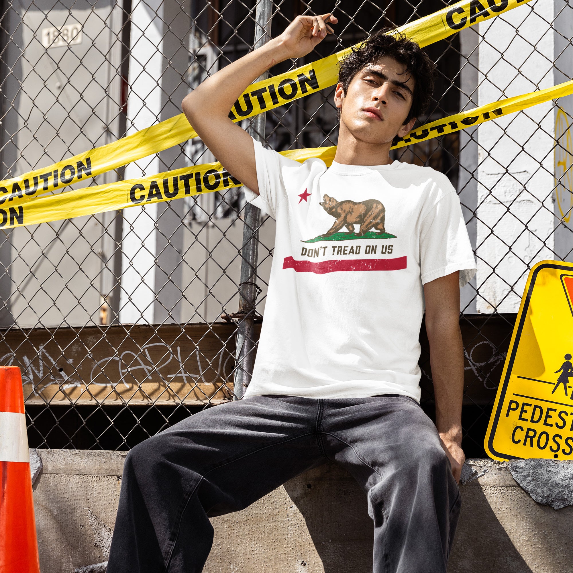 Young man leans against a fence draped in caution tape, wearing the California Gadsden Flag Shirt featuring a bear graphic and DON’T TREAD ON US slogan, symbolizing bold state pride and protest.