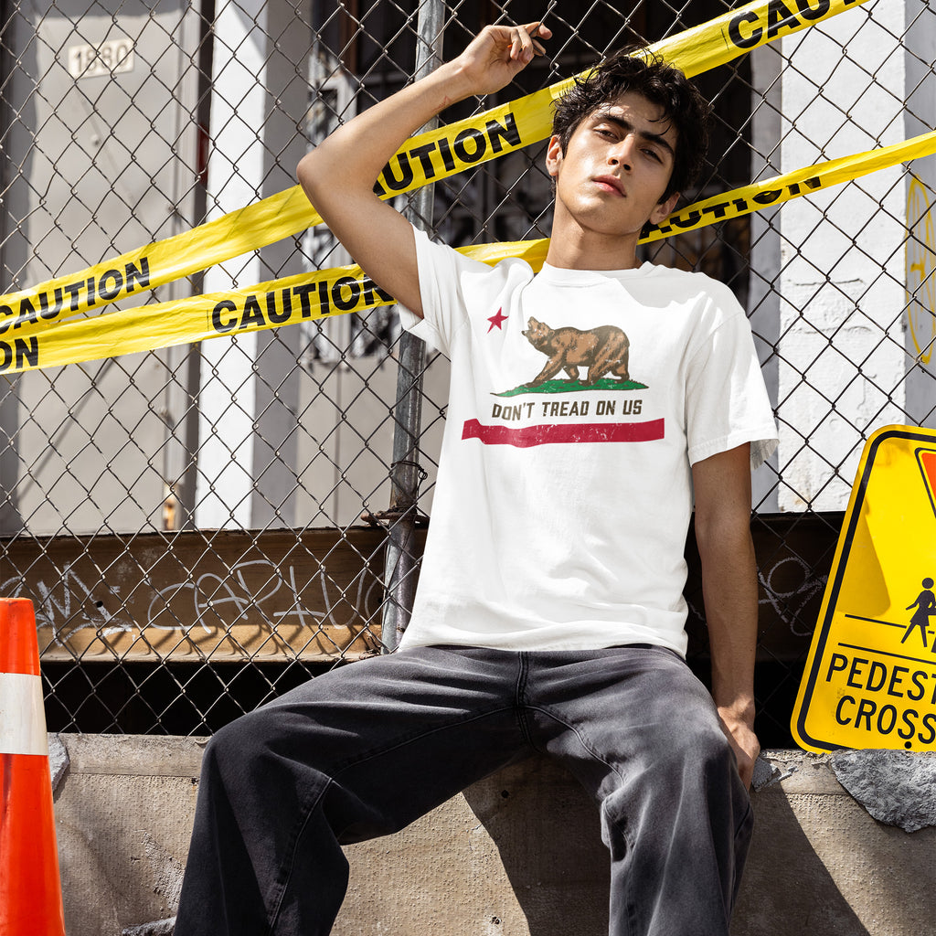 Young man leans against a fence draped in caution tape, wearing the California Gadsden Flag Shirt featuring a bear graphic and DON’T TREAD ON US slogan, symbolizing bold state pride and protest.