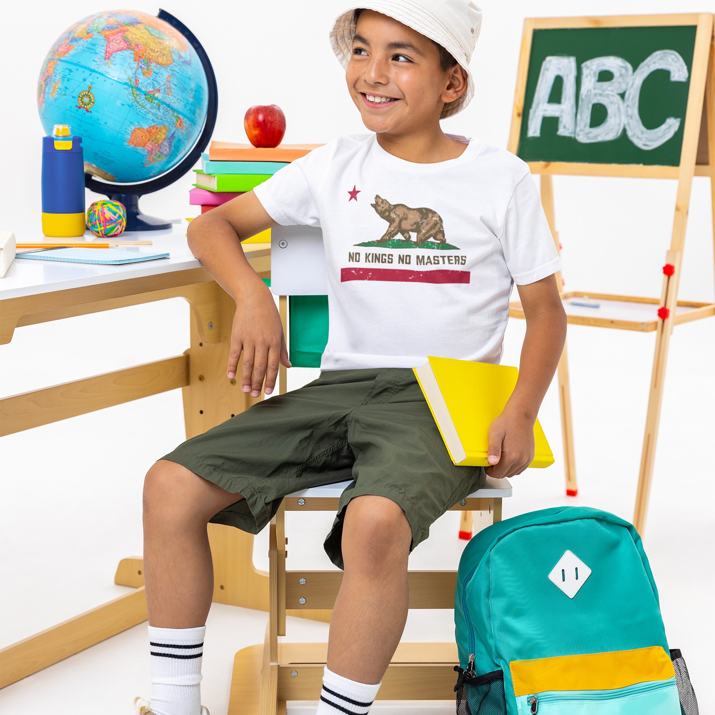 Young boy in a California No Kings Flag Shirt, showcasing a bear graphic and slogan, sits in a classroom setting, embodying rebellion and autonomy. He's surrounded by school items and a backpack.