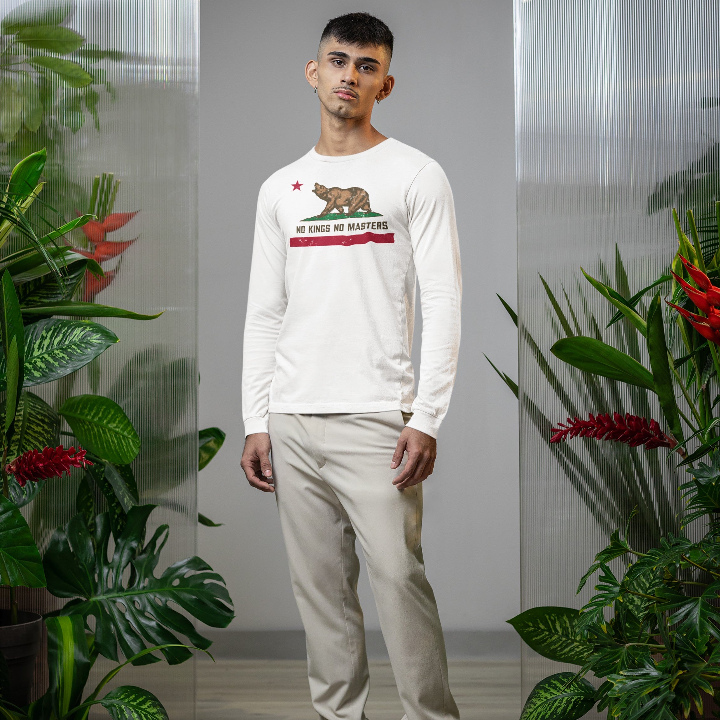 A young man models the California No Kings Flag Shirt, featuring a roaring bear and red star, amid lush tropical plants, highlighting its bold message of autonomy and power to the people.