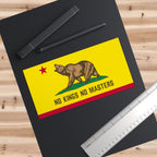 California Flag No Kings Sticker & Magnet features a brown bear on a yellow card with NO KINGS NO MASTERS, symbolizing resistance. Accompanied by pens and a ruler on a desk.
