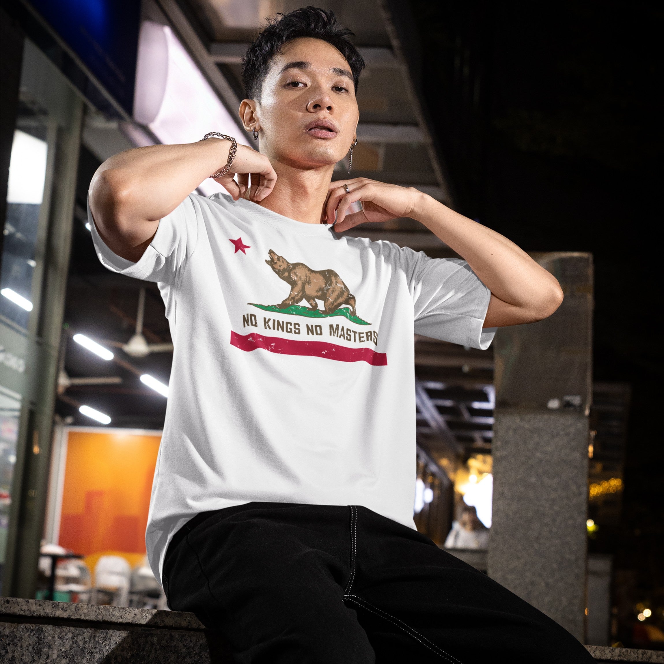 Young man in urban setting wearing California No Kings Flag Shirt, featuring a roaring bear and NO KINGS NO MASTERS slogan, symbolizing rebellion and autonomy. The design conveys a bold statement of independence.