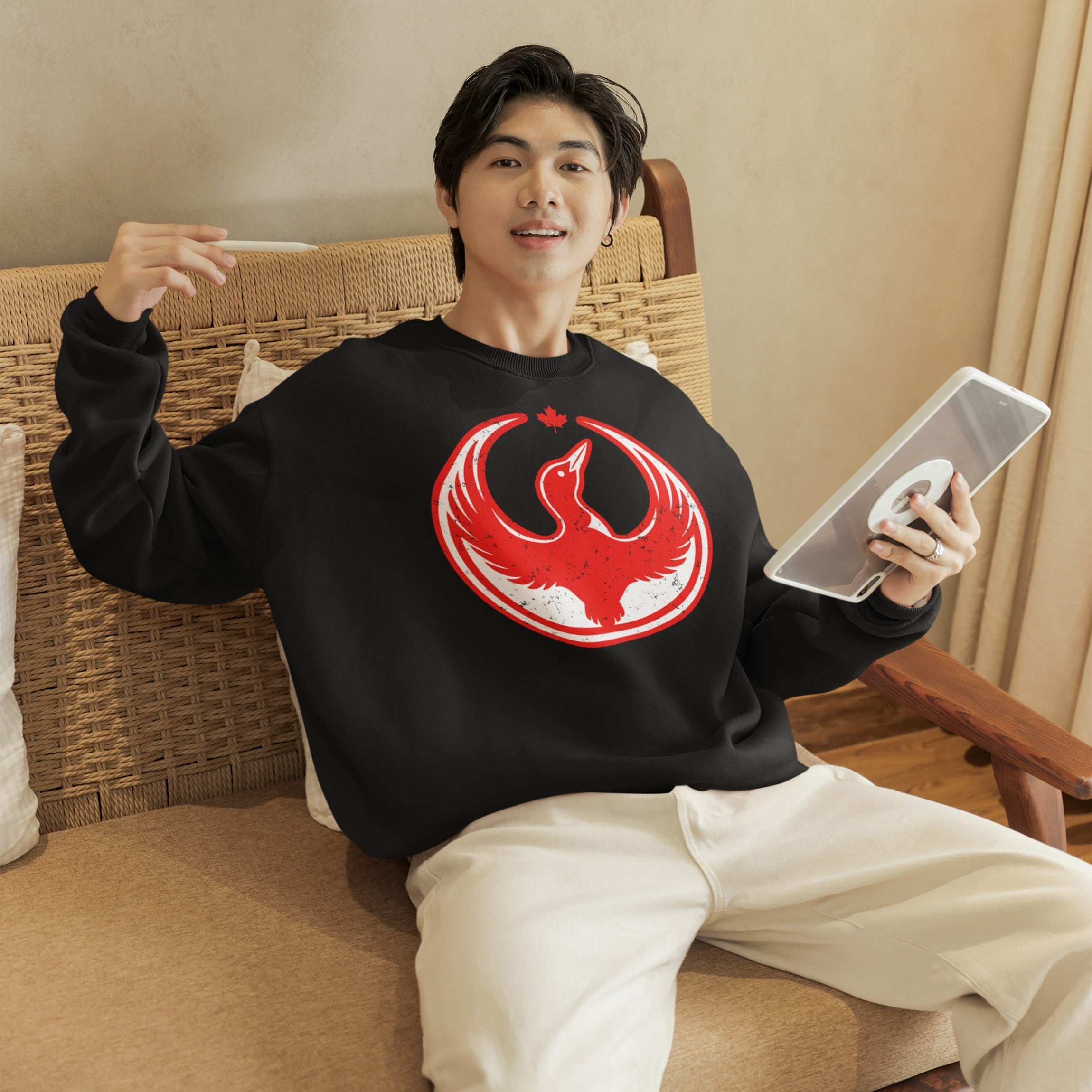 Young man reclines on a woven settee wearing a Canada Rebel Alliance crewneck sweatshirt, featuring a red-and-white phoenix emblem beneath a maple leaf, holding a stylus and tablet.