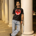 Young man at night wearing a Canada Rebel Alliance T-shirt with a red bird emblem, leaning against a column, showcasing a design symbolizing anti-fascist protest and support for equality.