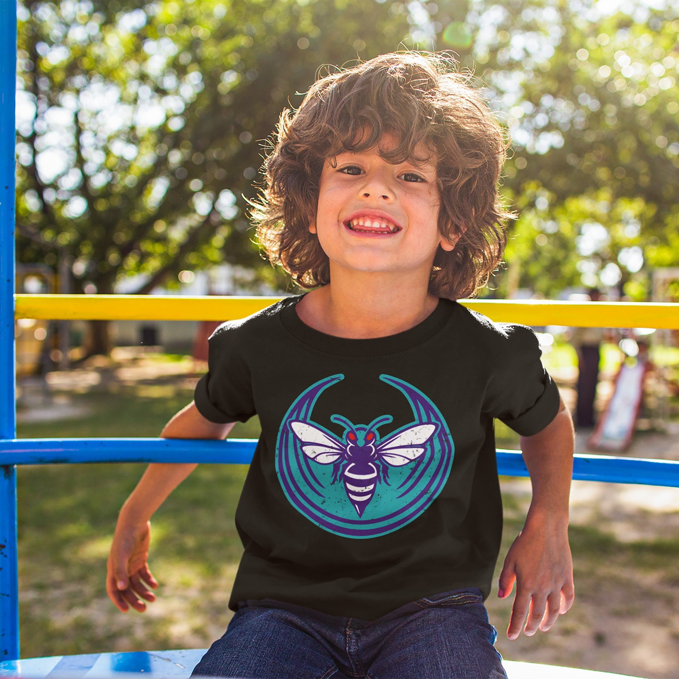 Young boy on playground bars wears a Charlotte Rebel Alliance T-shirt featuring a stylized hornet emblem, embodying anti-fascist and pro-equality values. The black tee is part of the inclusive collection.