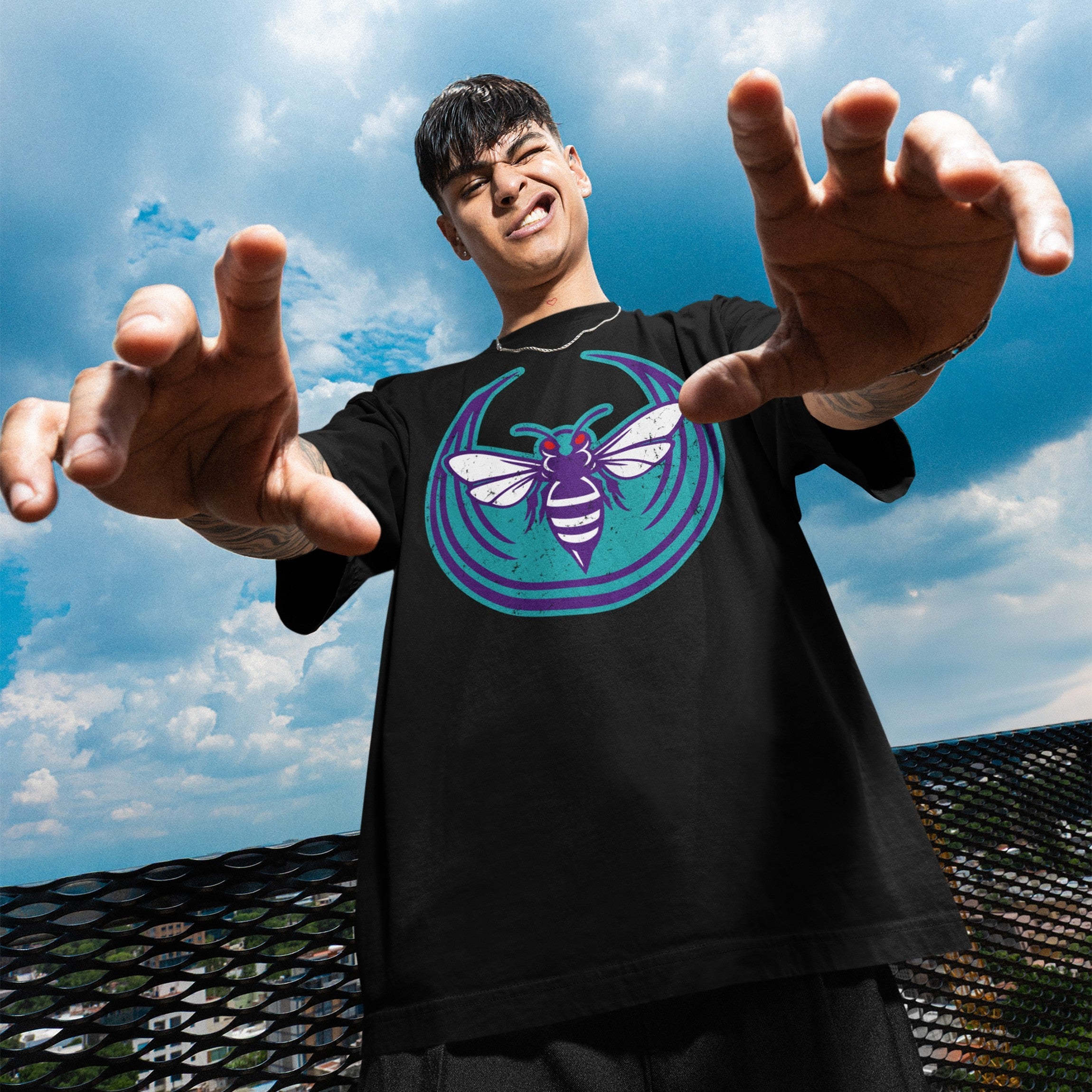 Young person on a rooftop wearing the Charlotte Rebel Alliance T-shirt, featuring a bold teal and purple hornet graphic, embodying anti-fascist, pro-equality messages. Their arms reach dynamically toward the camera.