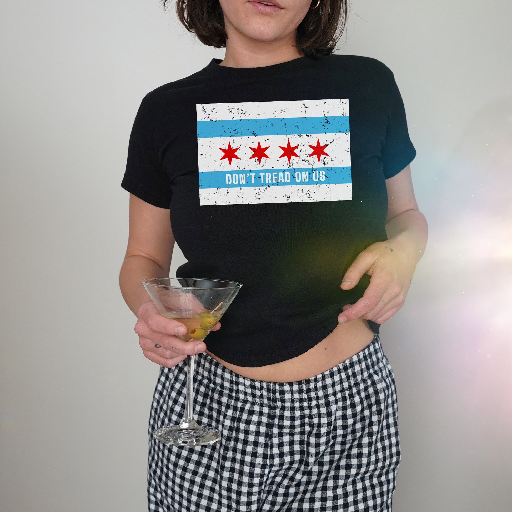 Chicago Gadsden Flag Women's Baby Tee featuring a cropped design with Chicago flag-inspired protest print, held by a person with gingham bottoms and a martini glass, embodying bold, stylish activism.