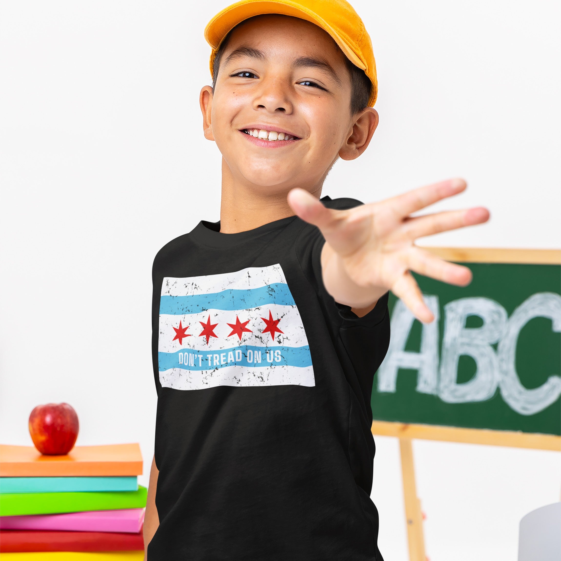 A school-age boy wears the Chicago Gadsden Flag Shirt featuring a stylized flag with “DON’T TREAD ON US,” symbolizing resistance. He stands playfully, reaching out in front of a chalkboard.