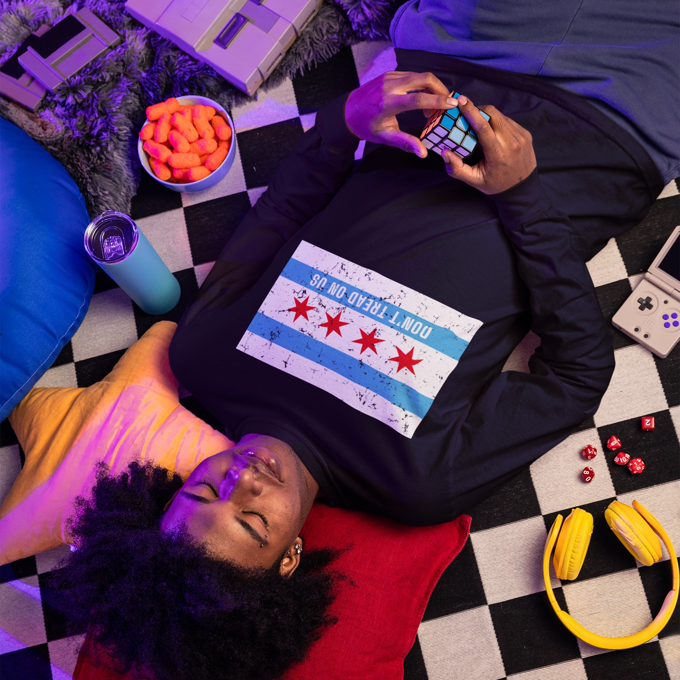 Young person on checkered mat wearing Chicago Gadsden Flag Shirt, showcasing a bold protest message, surrounded by retro gaming devices, dice, and snacks, embodying a relaxed, defiant spirit.