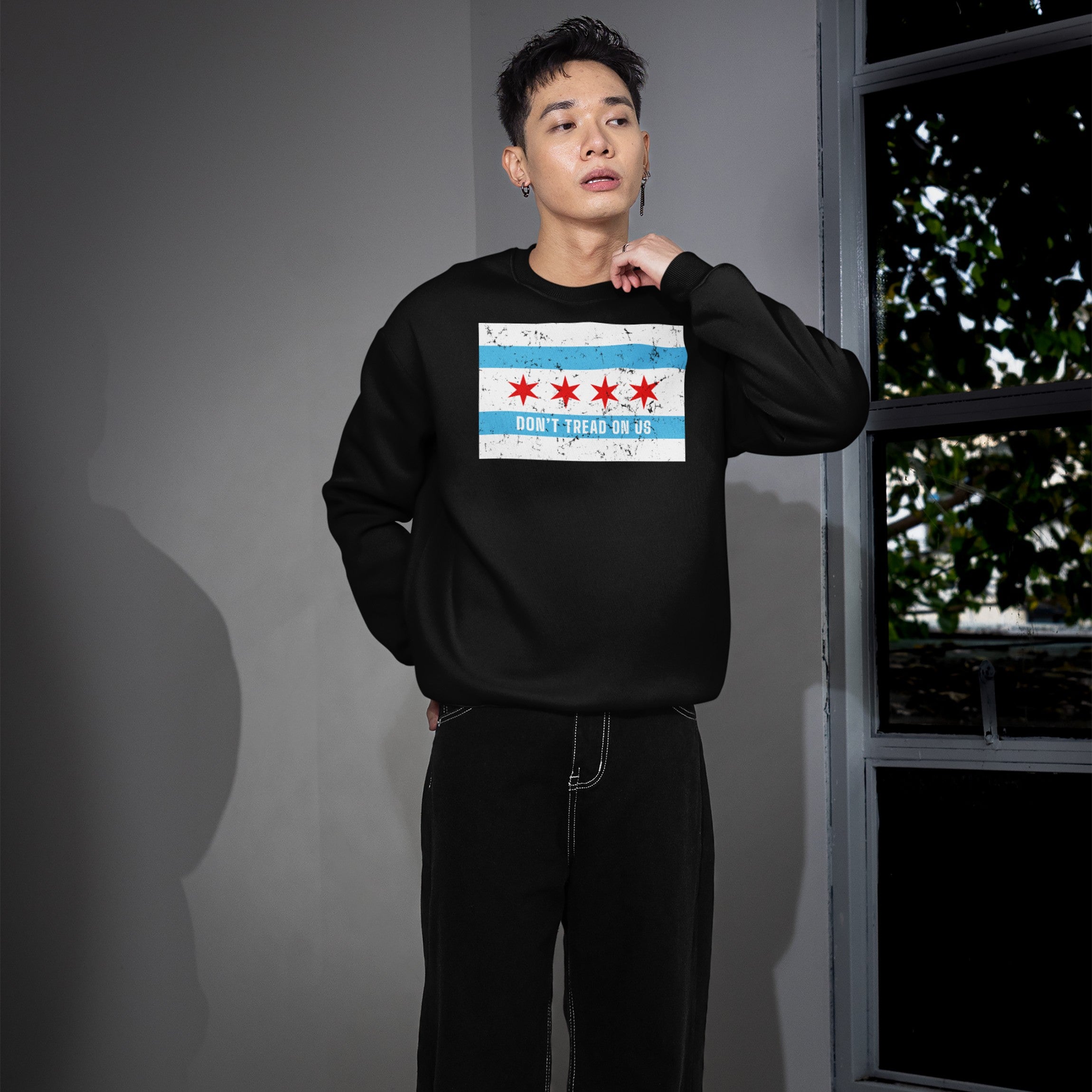 Young man indoors wearing a Chicago Gadsden Flag Shirt with a graphic of the Chicago flag and DON'T TREAD ON US slogan, embodying resistance and community spirit. He pairs it with loose black pants.