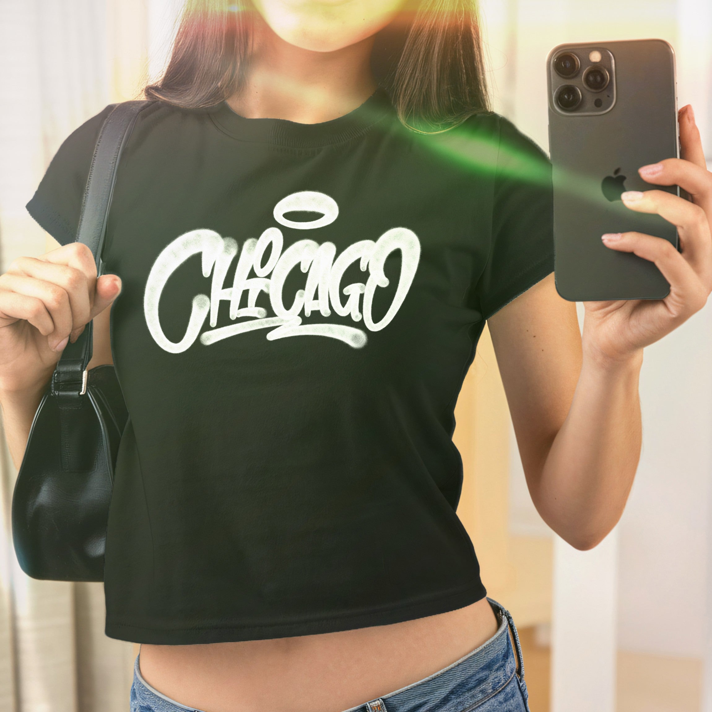 Mirror selfie showcasing the Chicago Handstyle Women's Baby Tee, featuring a graffiti-style CHICAGO print with a halo, worn with a black shoulder bag, embodying a retro Y2K aesthetic.