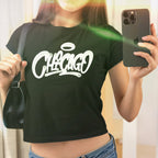 Mirror selfie showcasing the Chicago Handstyle Women's Baby Tee, featuring a graffiti-style CHICAGO print with a halo, worn with a black shoulder bag, embodying a retro Y2K aesthetic.