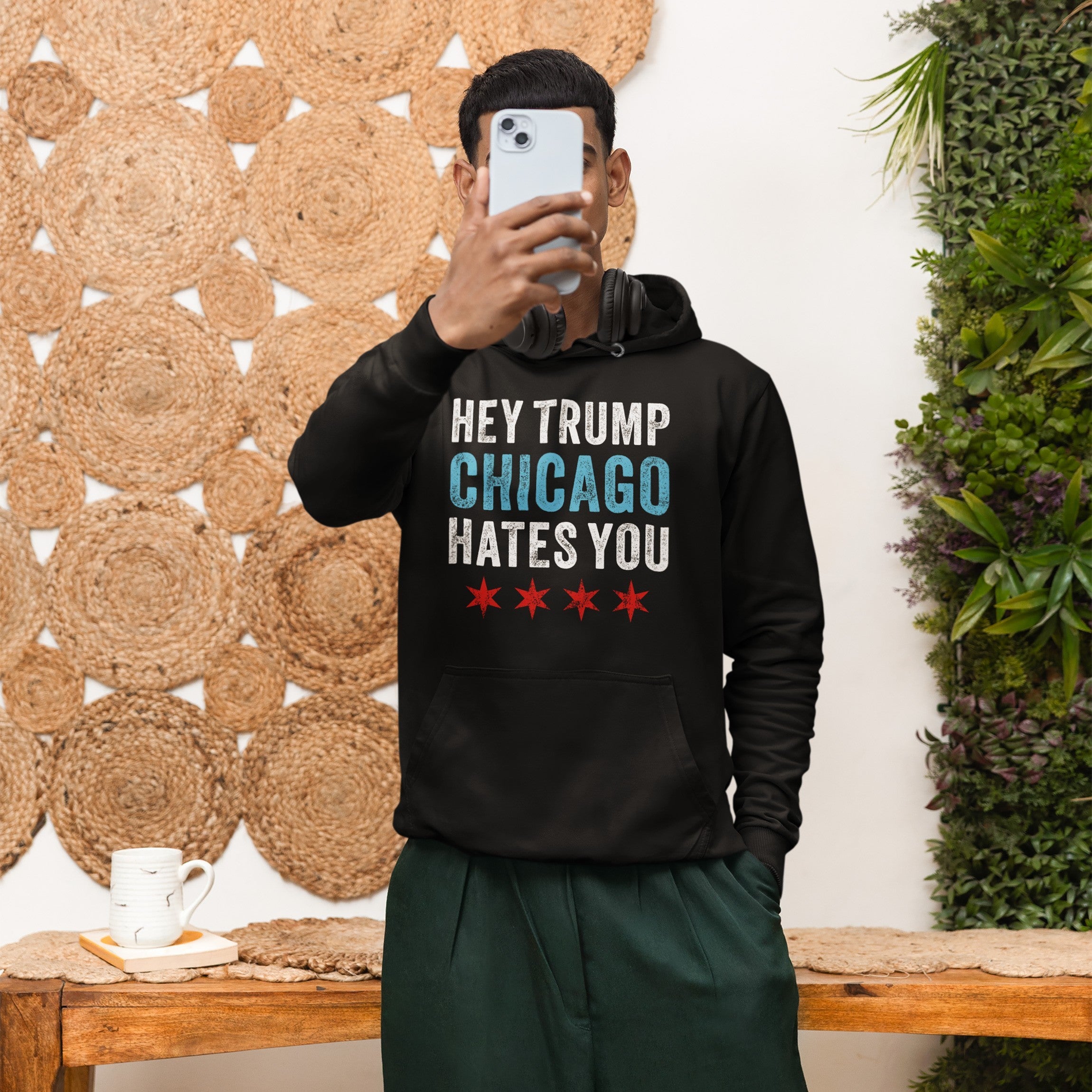 Person holds a smartphone wearing the Chicago Hates Trump hoodie with bold lettering and stars. Over-ear headphones rest around their neck, embodying protest spirit against a backdrop of woven mats.