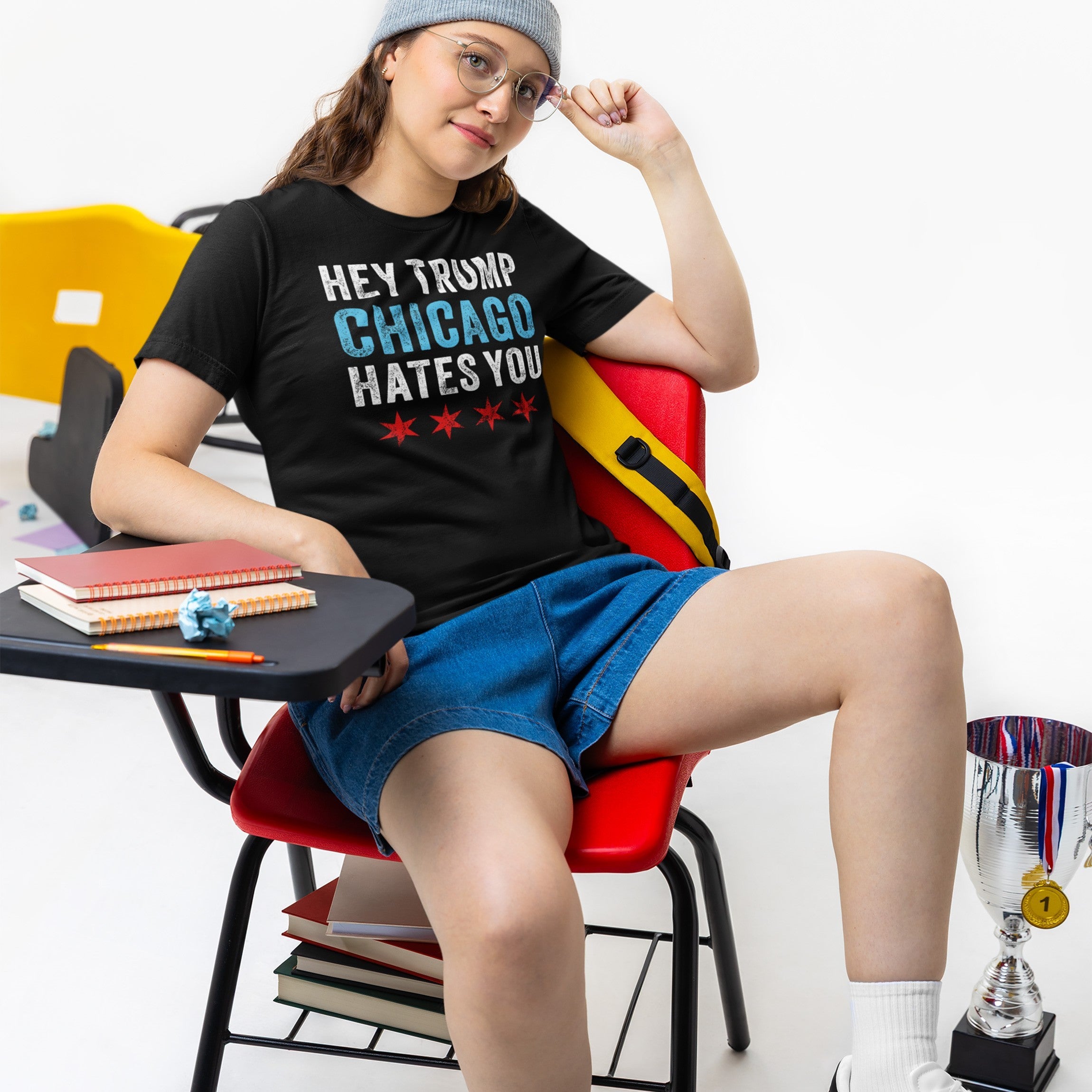A young person sits in a school-style chair wearing a Chicago Hates Trump T-shirt, conveying an anti-MAGA message with Chicago flag stars; a bold statement piece from a socially conscious brand.