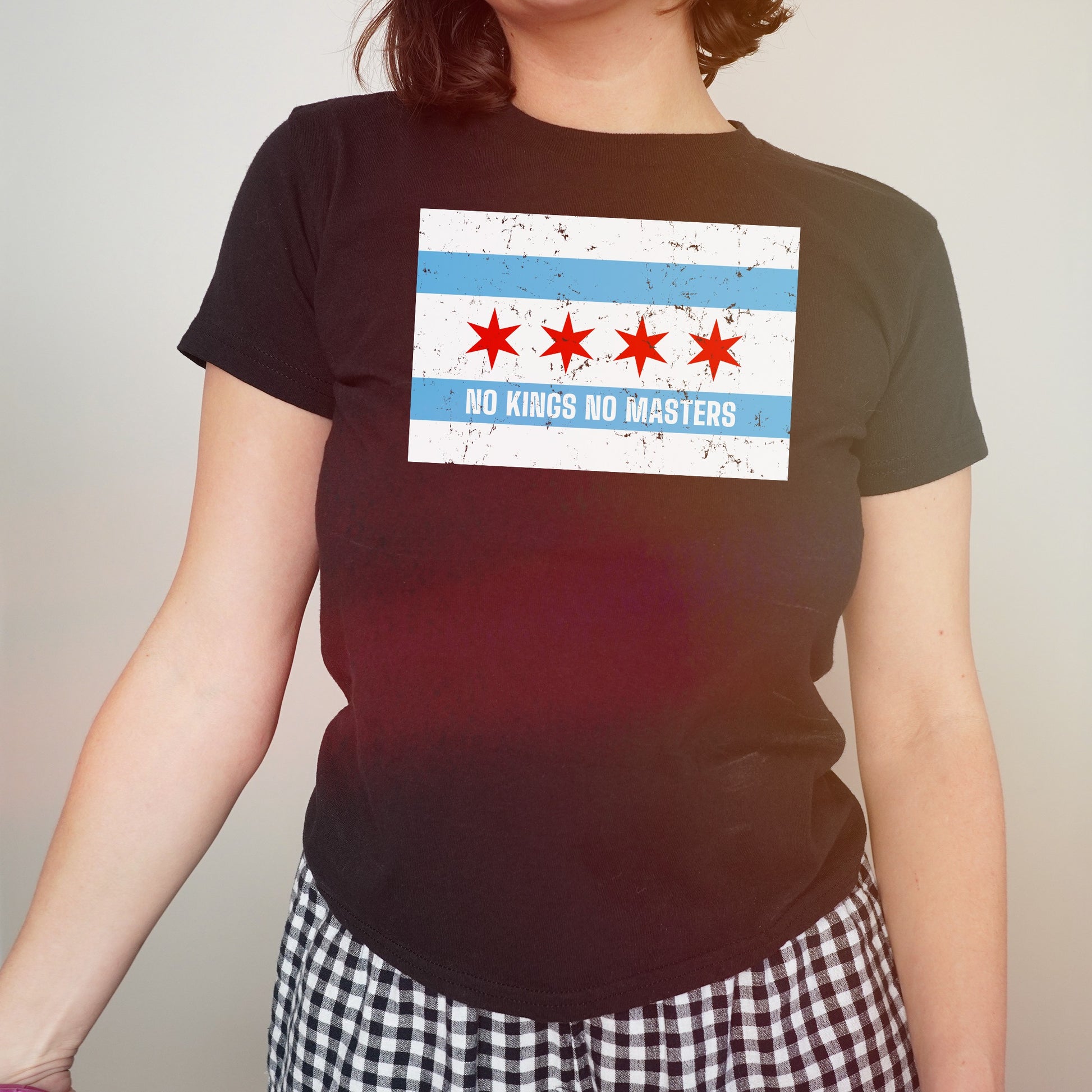 Chicago No Kings Flag Women's Baby Tee features a distressed city flag design with the slogan “NO KINGS NO MASTERS,” embodying resistance and freedom. Worn with black-and-white checkered bottoms.