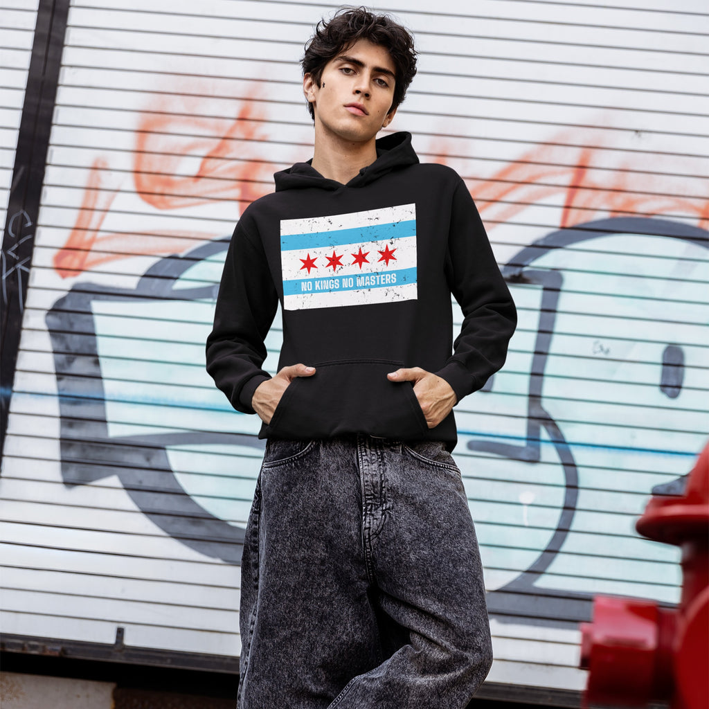 Young man in black Chicago No Kings Flag Shirt, featuring Chicago's flag and NO KINGS NO MASTERS slogan, leans against a graffiti backdrop, embodying urban rebellion and anti-authoritarian spirit.