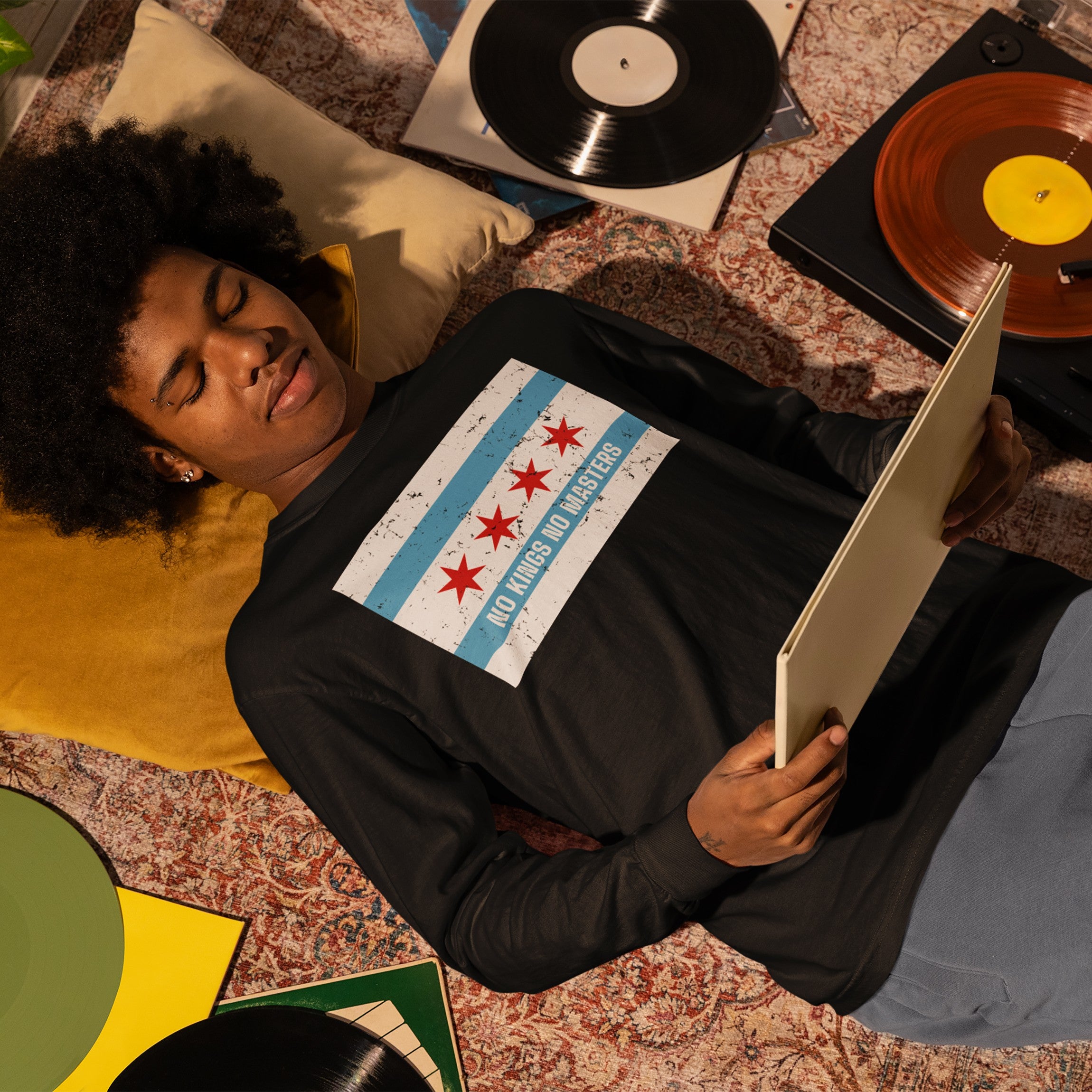 Person wearing the Chicago No Kings Flag Shirt, featuring Chicago's flag and NO KINGS NO MASTERS, lies on a patterned rug surrounded by vinyl records and a turntable, embracing freedom and equality.