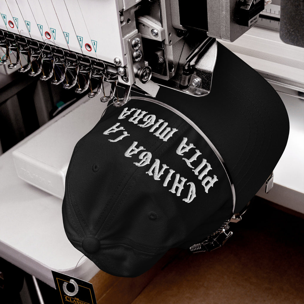 A black Chinga La Migra Hat in an embroidery machine, with white thread stitching an anti-fascist Spanish slogan on the front, embodying bold protest against immigration enforcement.