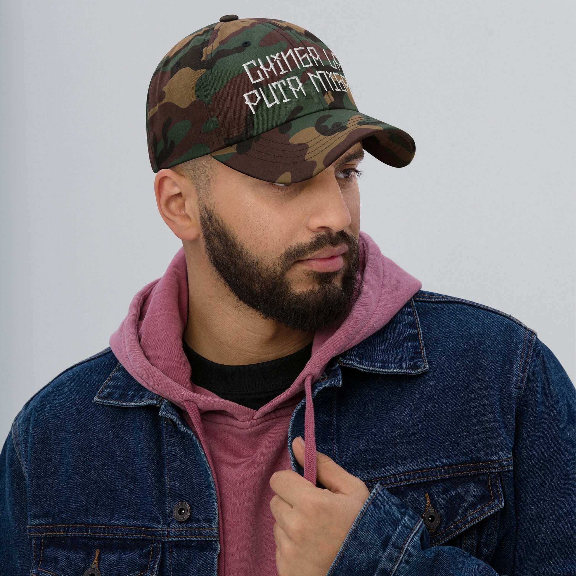 A man wears the Chinga La Migra Hat, a camouflage-print cap with bold white embroidery, conveying a strong message against immigration enforcement, paired with a mauve hoodie and denim jacket.