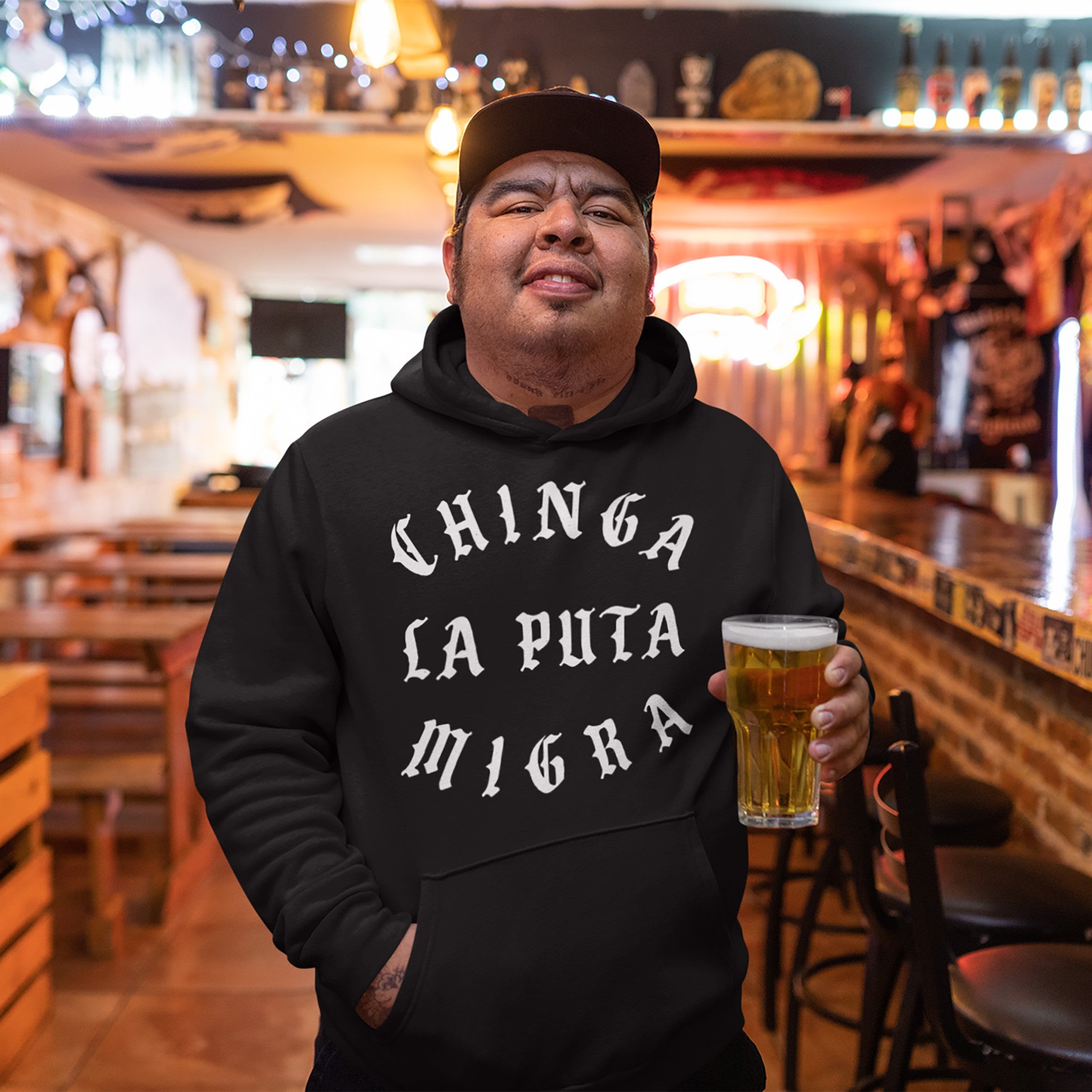 Man at bar wearing Chinga La Migra Hoodie, featuring Chinga La Puta Migra in white Gothic letters. Holds beer pint amid casual setting with wooden furniture, embodying protest spirit and immigrant rights advocacy.