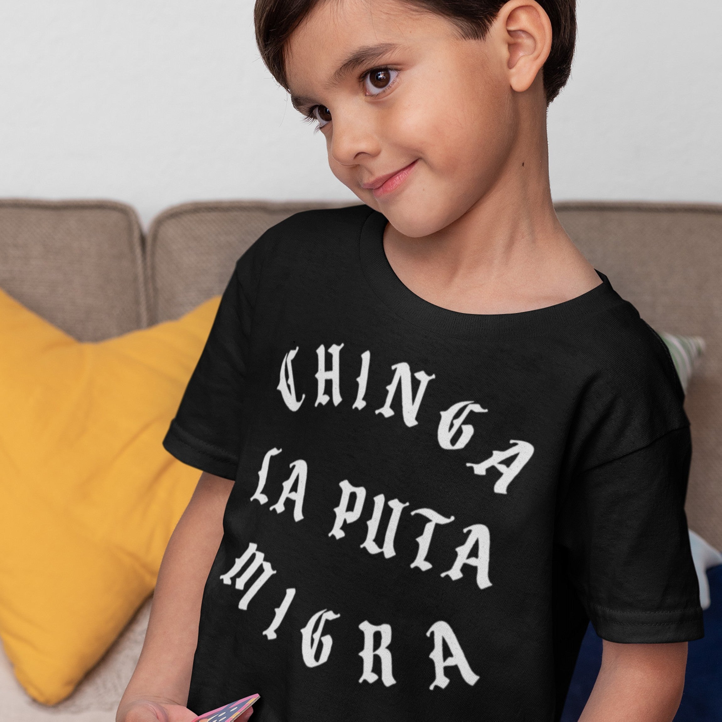 Young boy on couch wears the Chinga La Migra T-shirt, featuring bold, white Spanish text in vintage blackletter type, ideal for anti-fascist and immigrant rights supporters.