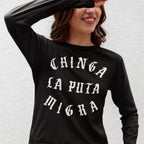 Young woman in a black Chinga La Migra T-shirt, featuring bold, Gothic-style white text promoting anti-fascist messages. She shields her eyes playfully, embodying a spirit of protest and unity.