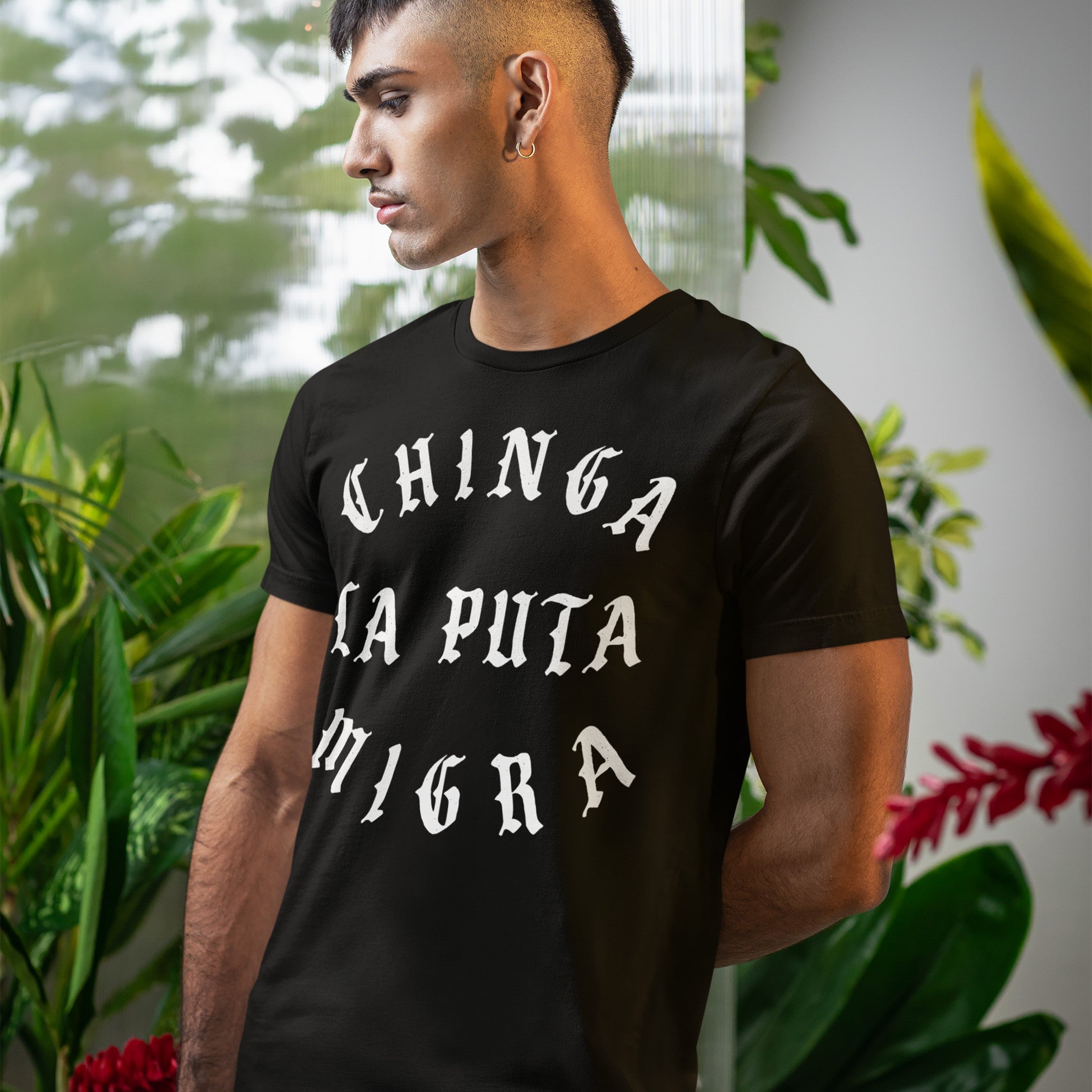 Young man wearing a Chinga La Migra T-shirt with bold white Old English lettering, standing indoors near tropical plants, capturing an anti-fascist message in a slightly distressed blackletter style.