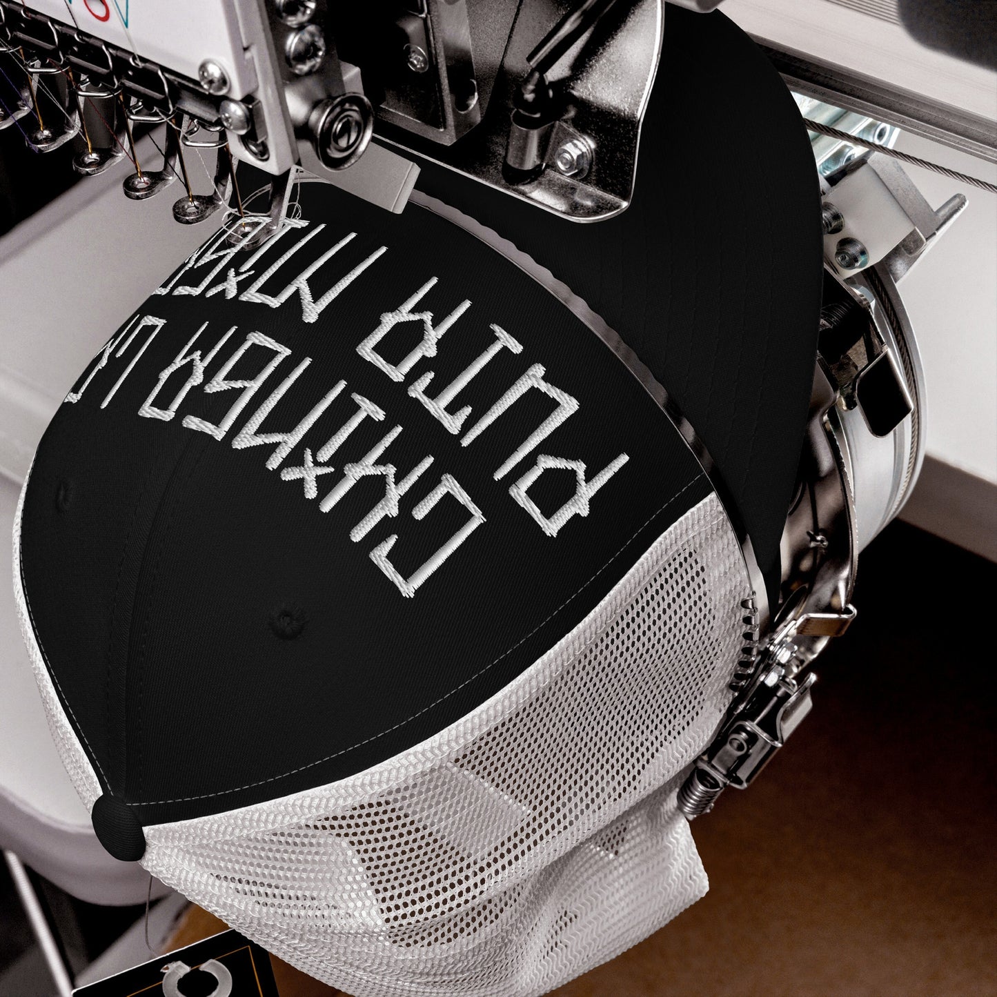 Close-up of Chinga La Migra Hat being embroidered on an industrial machine, showcasing the bold block-style lettering on a black front panel with white mesh back, symbolizing resistance against oppression.