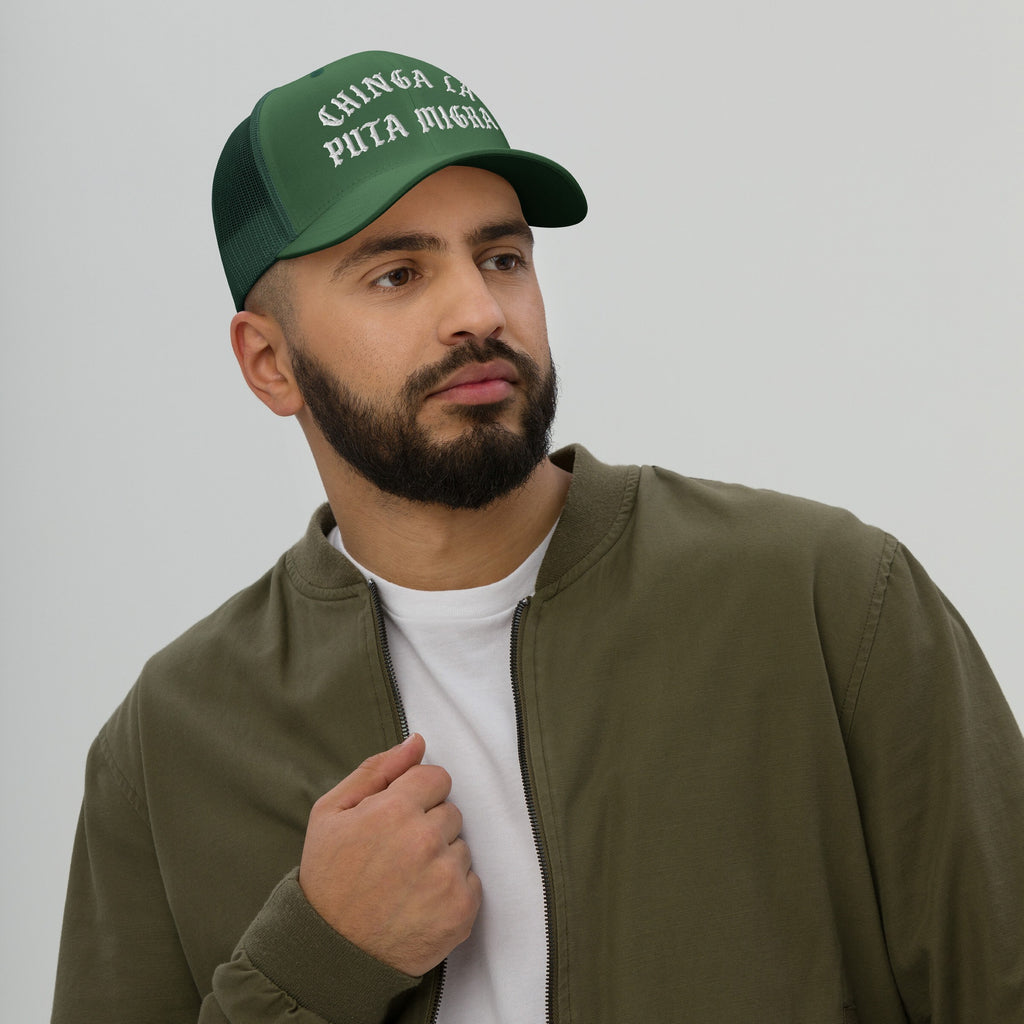 Bearded man wearing 'Chinga La Migra Hat' in dark-green with white Old English embroidery, expressing an anti-fascist message. His casual attire complements the protest-themed trucker cap's bold statement.