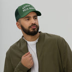 Bearded man wearing 'Chinga La Migra Hat' in dark-green with white Old English embroidery, expressing an anti-fascist message. His casual attire complements the protest-themed trucker cap's bold statement.