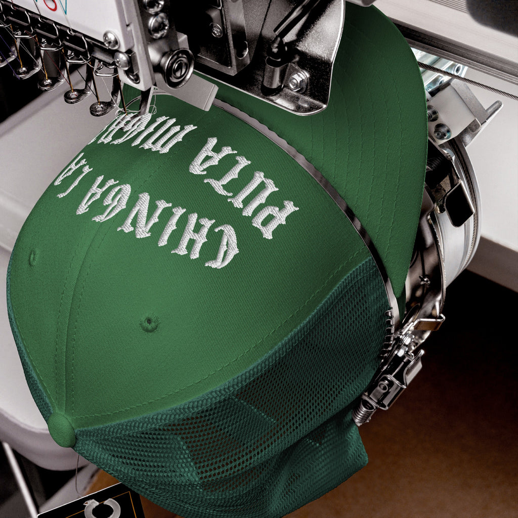 Green mesh-back Chinga La Migra Hat in embroidery machine, white thread stitching bold protest message on front, encapsulating defiance against ICE, highlighting immigrant rights and anti-fascist stance.