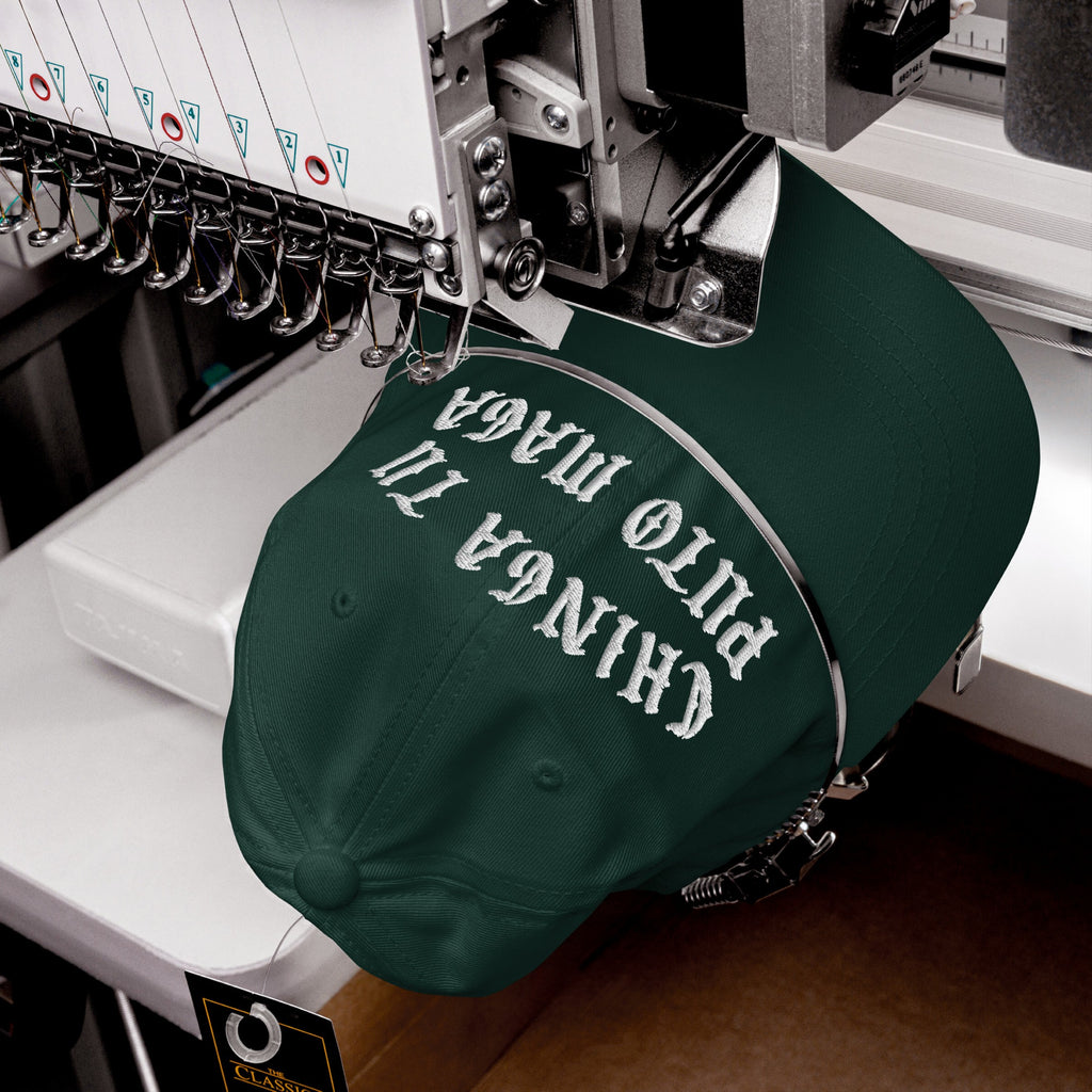 Chinga Tu MAGA Hat on embroidery machine, dark-green cap with bold white lettering, showcasing anti-fascist message in Spanish. Hat clamped for stitching, multi-needle head visible.