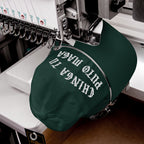 Chinga Tu MAGA Hat on embroidery machine, dark-green cap with bold white lettering, showcasing anti-fascist message in Spanish. Hat clamped for stitching, multi-needle head visible.