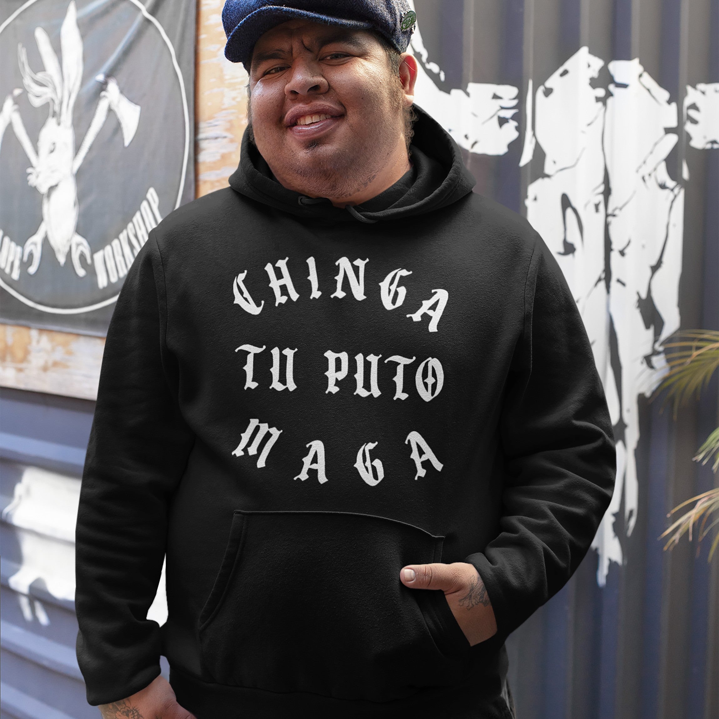 Man wearing the Chinga Tu MAGA Hoodie in a workshop, featuring bold, vintage-style blackletter text. The setting includes a sign with a rabbit and crossed axes, reflecting a defiant, anti-fascist theme.